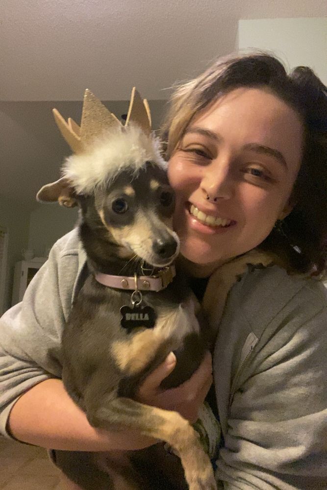 Me holding my chihuahua miniature pinscher mix, Bella. Bella is wearing a gold and white birthday crown and her purple collar. I am wearing a gray jacket. 