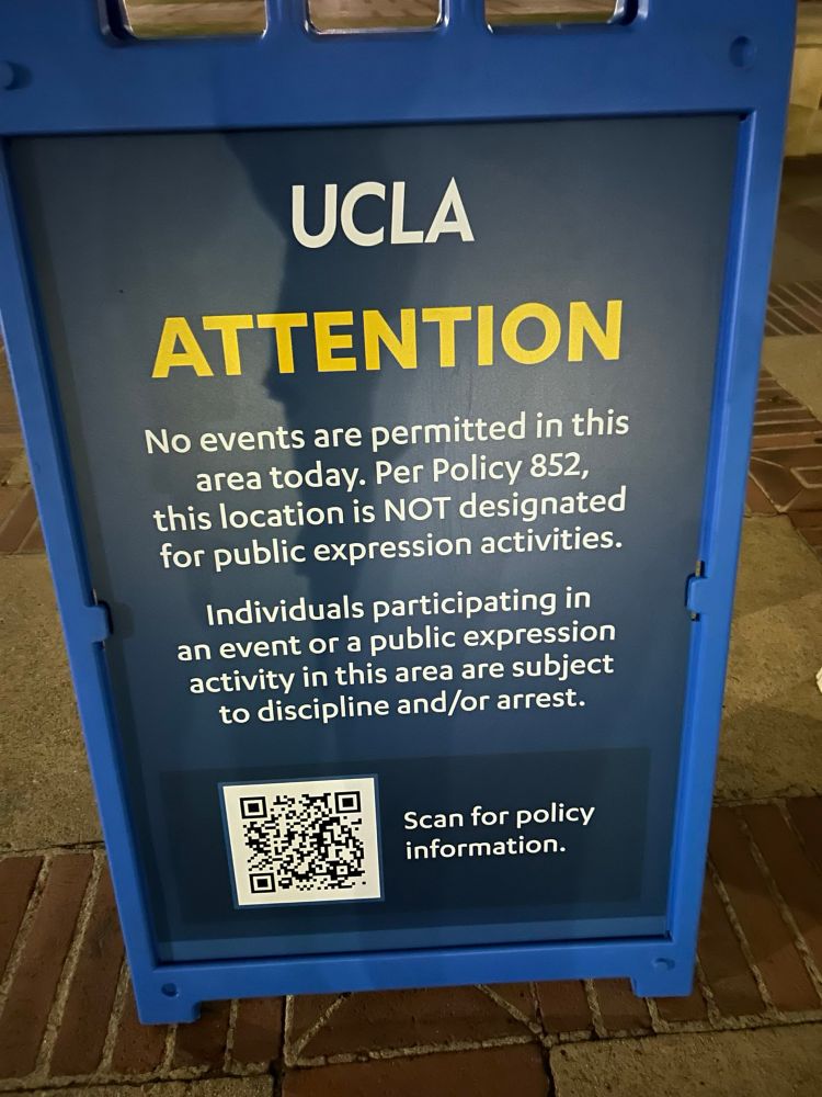 An easel sign reads, “UCLA
ATTENTION
No events are permitted in this area today. Per Policy 852, this location is NOT designated for public expression activities.
Individuals participating in an event or a public expression activity in this area are subject to discipline and/or arrest.
Scan for policy information.”
