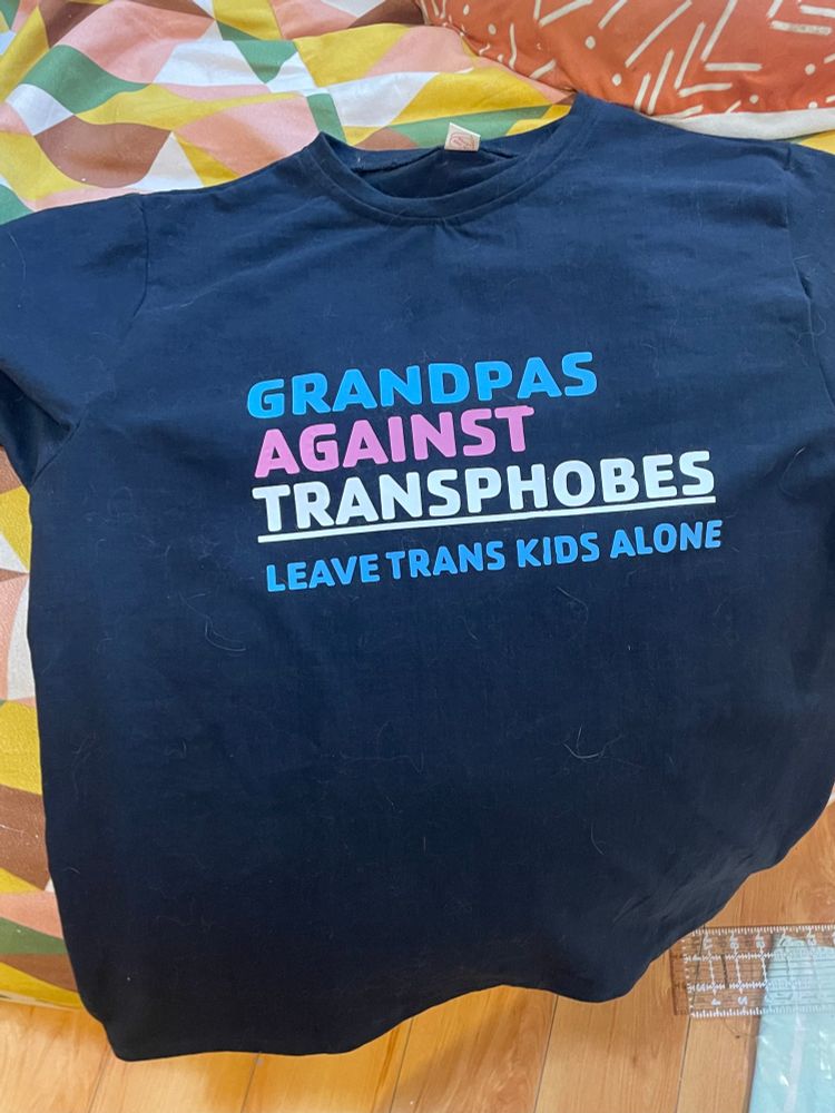 Black t-shirt reads GRANDPAS AGAINST TRANSPHOBES / LEAVE TRANS KIDS ALONE, in blue, pink, and white.