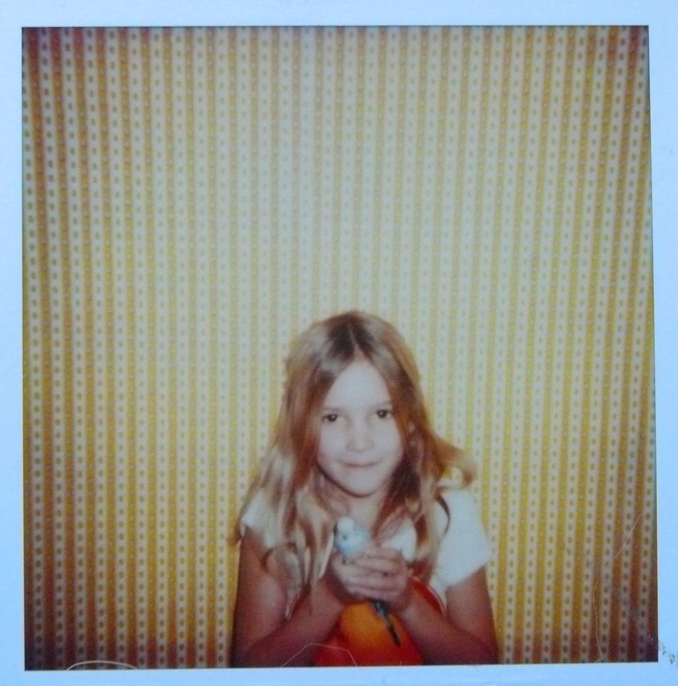 Young female presenting child about 7 years old holding a blue parakeet in her hands and smiling. Looks a bit ominous, but she did love the bird with her heart and soul. Wearing a white t-shirt with a sunset/rise? on it in front of yellow striped and patterned wallpaper.