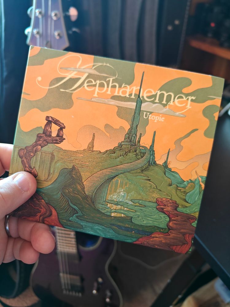 Aephanemer's new album Utopie, in digipack CD format. The cover is a painting of a distant tower across a fantasy landscape. 