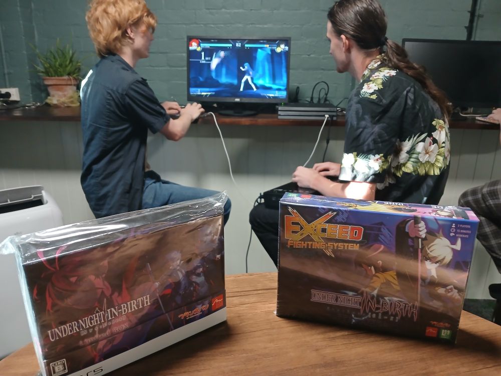 The background shows a screen displaying the video game, Melty Blood Type Lumina, in the center of the image. Two players are seated side by side, facing the screen with controllers in their hand. 
In the foreground, there are two boxes, side by side. The left box is a collector's edition of Under Night In-Birth 2 for the Playstation 5 in its original plastic packaging. The right box contains a copy of the Under Night board game, also it its original plastic packaging. 
