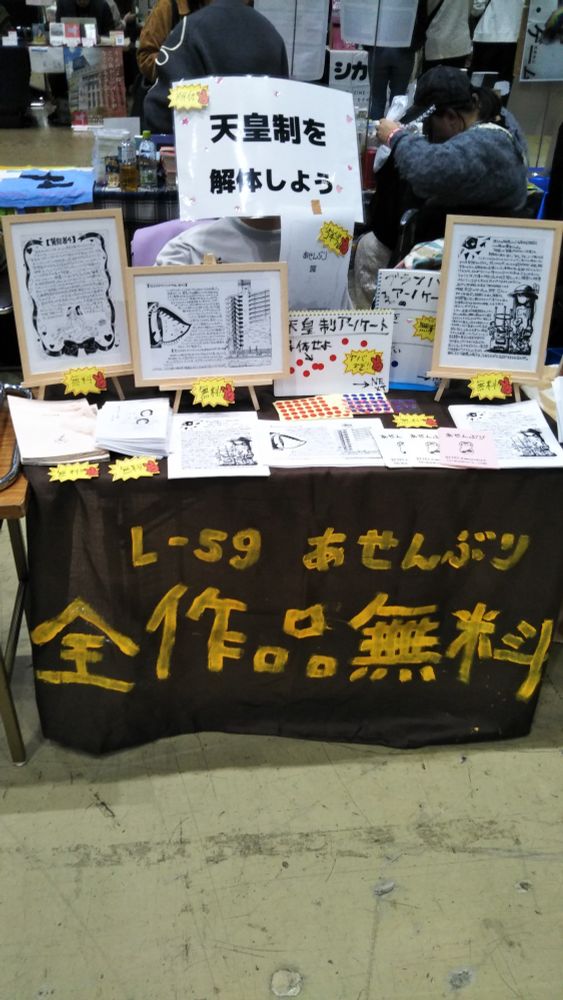 東京文フリ会場での「あせんぶり」のブースの写真。「全作品無料」と書かれた垂れ幕、「天皇制を解体しよう」と書かれたプラカード、額に入った作品が並べられている。