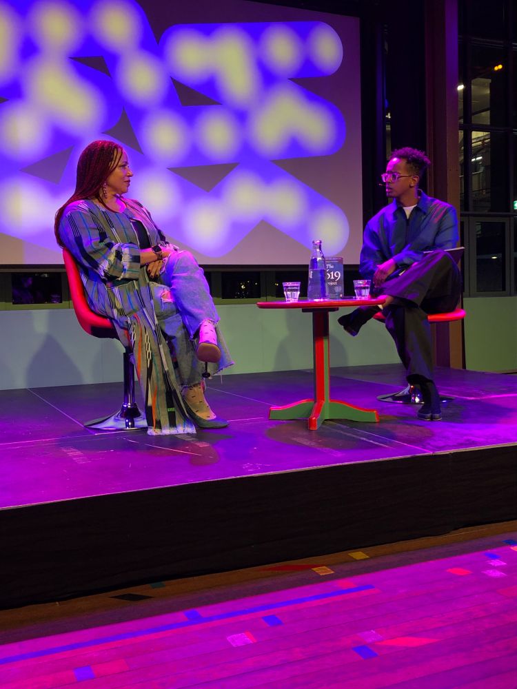Nikole Hannah-Jones seated on a chair on stage, next to a small side table, with Hani Saleh on the other side of the table. A water bottle, two glasses, and a copy of 1619 are on the table.