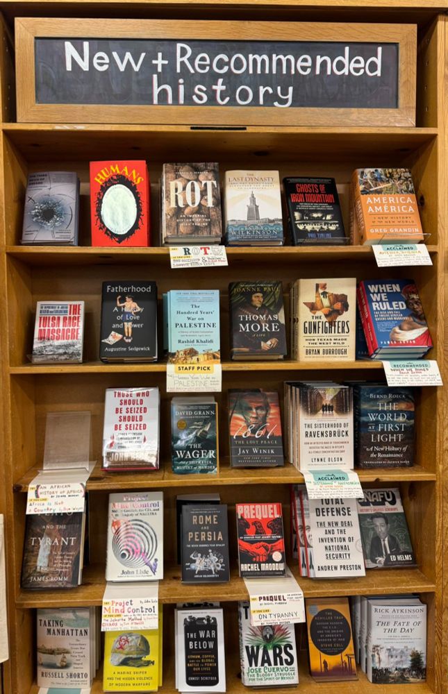 A narrow wooden bookcase with five shelves, and "New & Recommended History" on a chalkboard at the top. Each shelf holds 5-6 books. On the top shelf, second from the left, is a copy of HUMANS, with its silvery oval mirror, black frame, red background and yellow typeface.