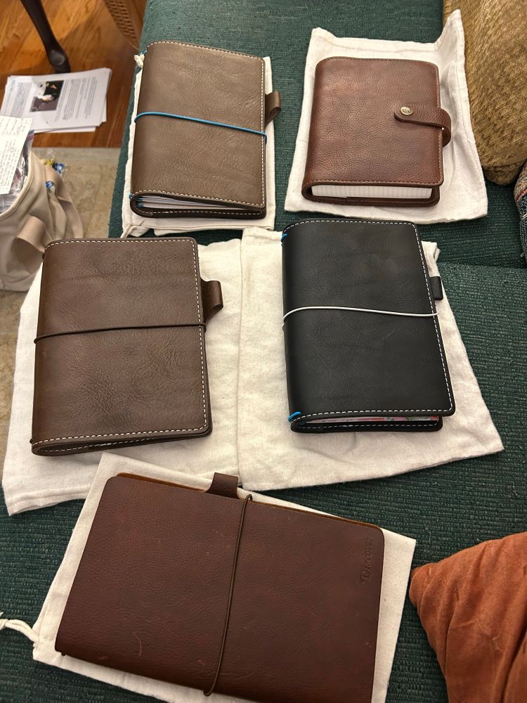 Current travelers notebook loot spread out on the couch next to me. In the foreground is a well patinaed Narrow or original Travelers Notebook sized cover with inserts from the Travelers Notebook company. Chic Sparrow Outlander Sojourn Tan. 
The next closest row has two filled notebooks, the one on the left a CS Chocolate Creme with white contrast stitching in B6 with inserts from Paper Penguin and ThreeTreeWhimsy. 
On the right is a CS Black Beauty Creme with bright blue elastics and white contrast stitching in B6 Slim. It also has a mix of inserts from Paper Penguin and ThreeTreeWhimsy. 
The furthest row has on the left a CS Chocolate Creme in Wide with white contrast stitching and what was meant to be turquoise elastic strings. It is loaded with Bookfell Studios inserts. To the right of it is a CS Strider in the notebook design Number 10. It has a snap closure and can work as a folio or a TN. Inside is a thick book like insert from CreateWithPen. 
