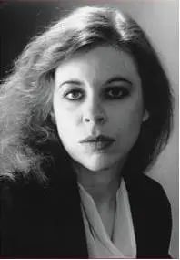Black and white photo portrait of British fantasy/horror author Tanith Lee. She is wearing a scoop-necked shell in white and a black blazer. Her facial expression is intense and intellectually formidable, eyes rimmed in kohl, her shoulder-length hair streaming to the side as if in a light breeze. An overhead light source makes her hair shine at the crown of her head. A brilliant and prolific author. 