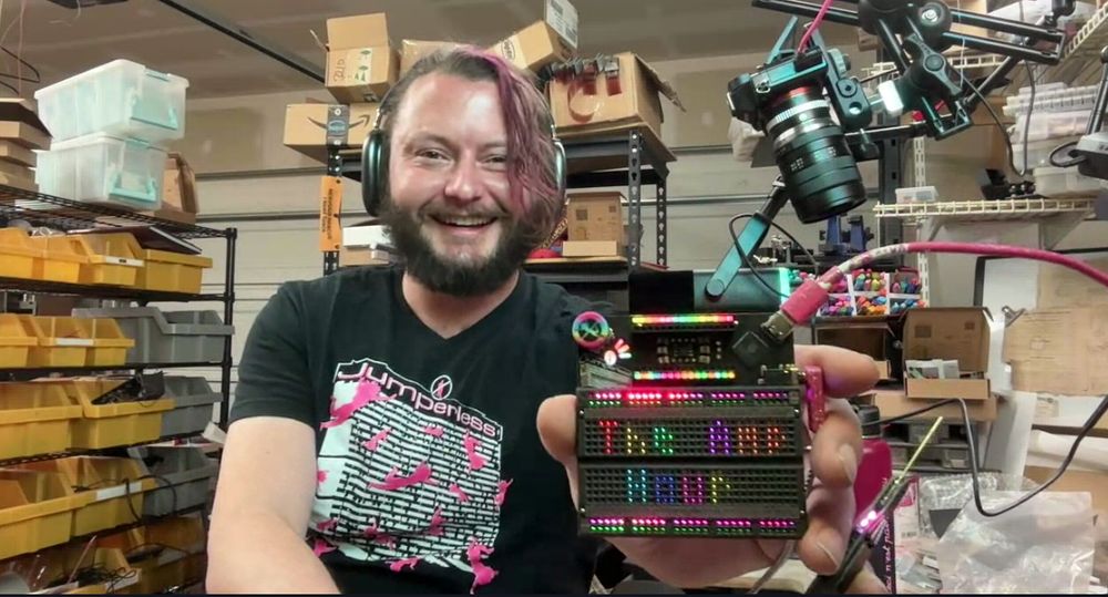 Kevin is holding up a Jumperless breadboard with the words "The Amp Hour: in the LEDs backlighting the holes. The board is plugged into a computer via USB and Kevin is in his garage lab.