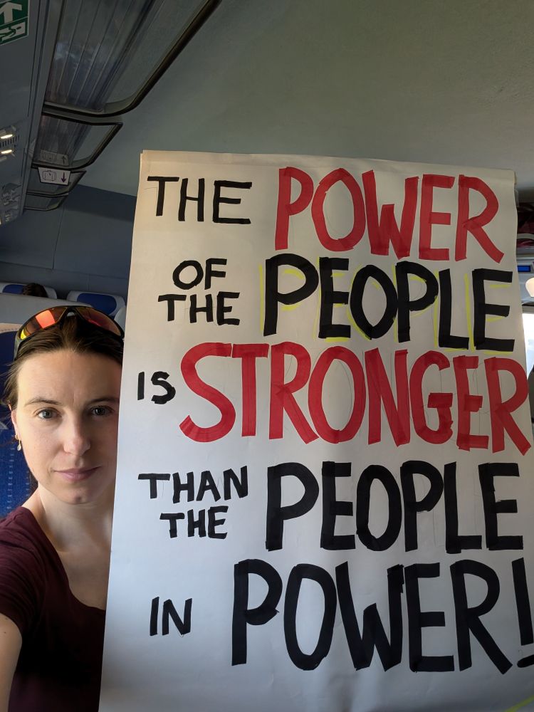 A picture of Emma, the post author, on a train holding a homemade sign stating "the power of the people is stronger than the people in power"