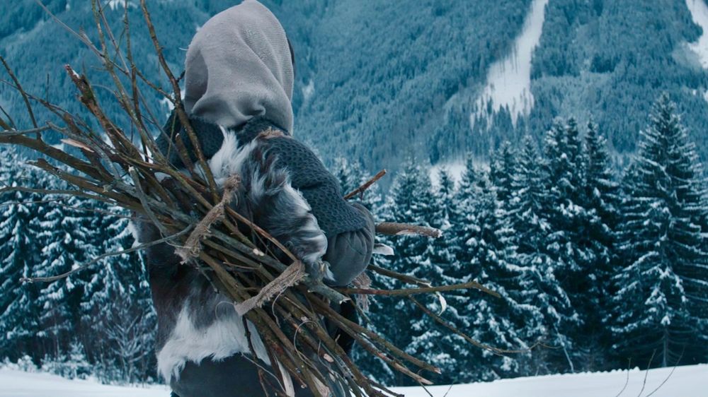 We see the back of a woman dressed in furs and warm fabrics as she looks out into a vast winter tundra. A bundle of bound sticks sits on her back. 