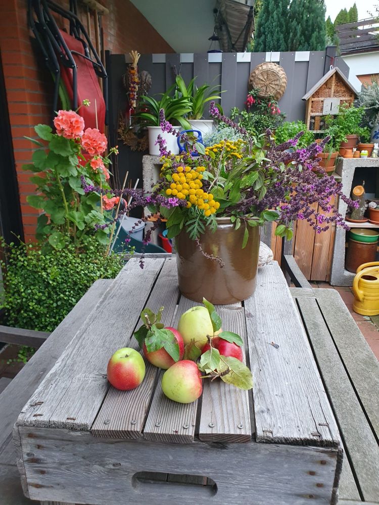 Ein Wiesenblumenstrauß in einer tönernen Vase steht auf einem alten, silbergrauen Holztisch. Daneben liegen ein paar pflückfrische Äpfel.