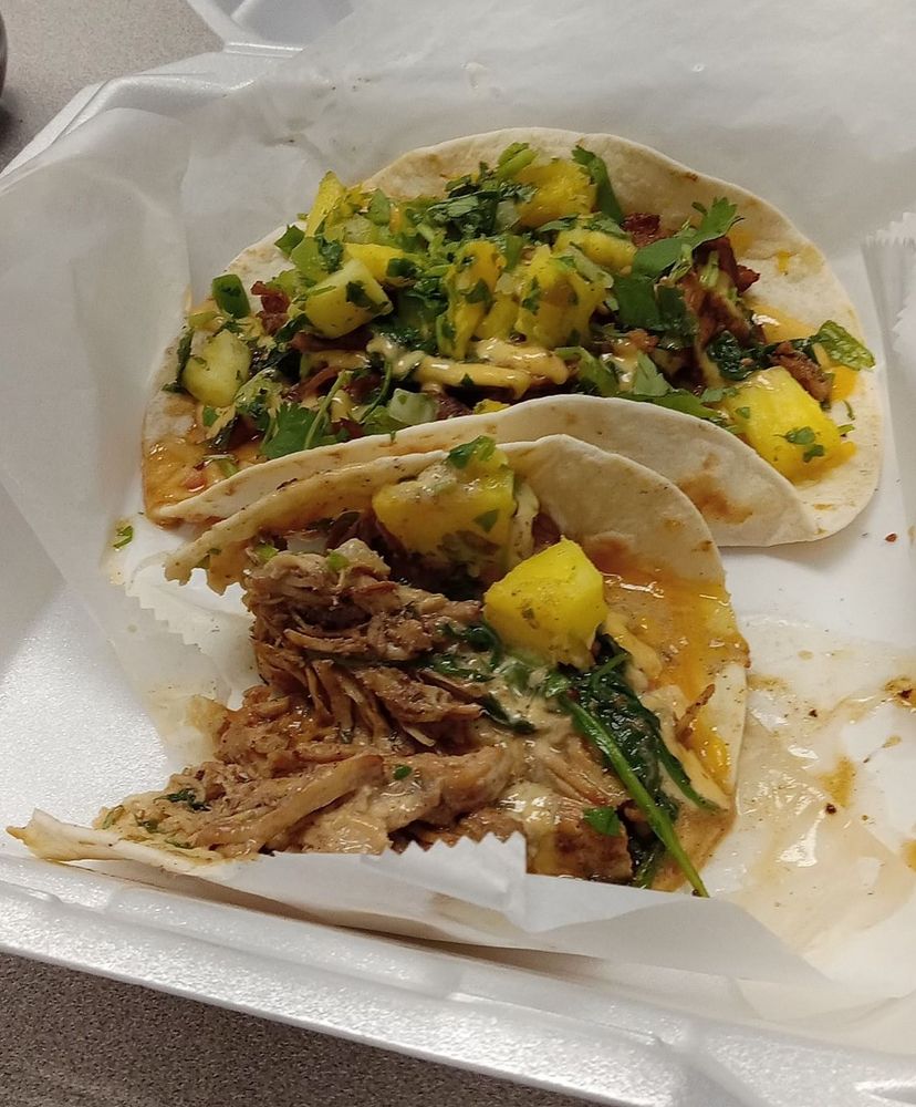 Two flour tortilla tacos in a foam serving container. They are made with shredded jerk chicken, cilantro, smoked habanero garlic sauce and pineapple salsa.