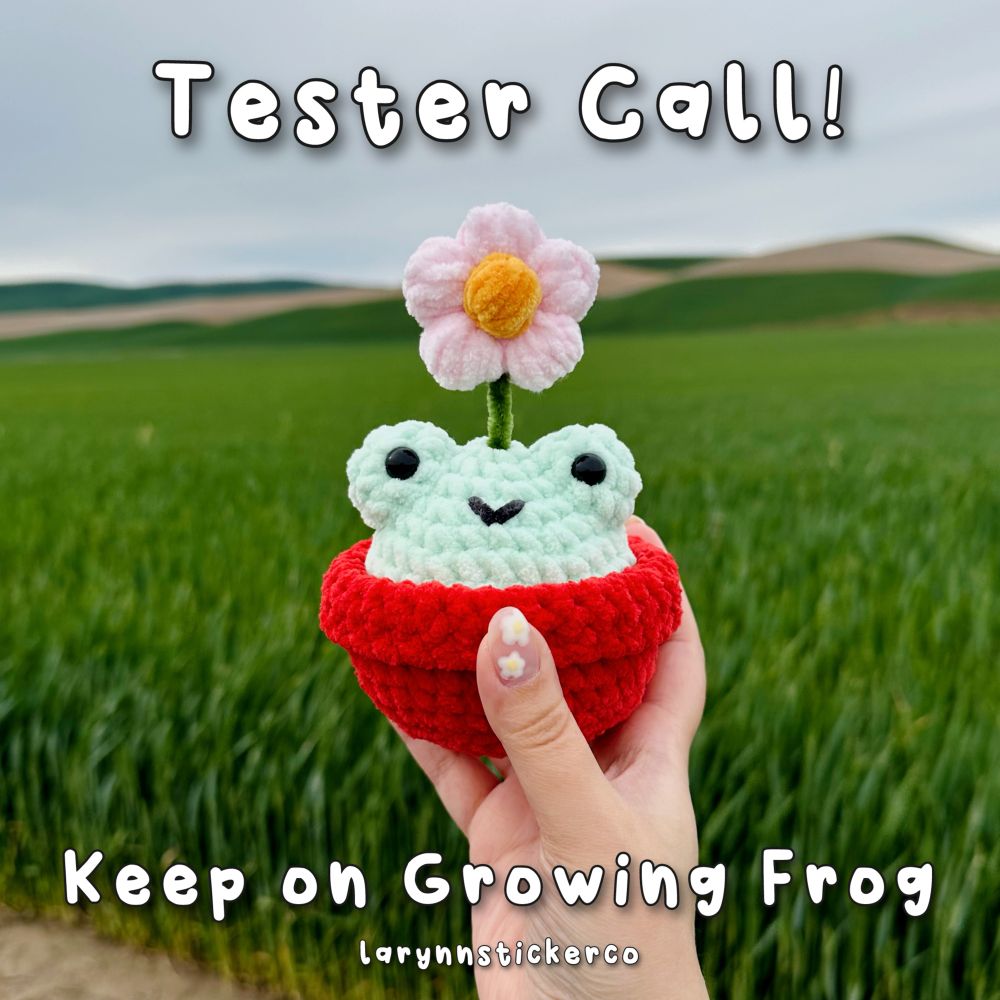 Text: Tester call! Keep on Growing Frog from LaRynn Sticker Co.

Held up in front of a blue sky and green hills is a crocheted frog. The green frog sits in a red pot, and has a pink flower coming out of the top of its head. 