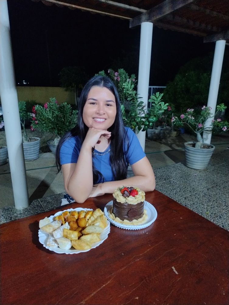 Eu sou uma mulher de pele morena clara, cabelos pretos lisos, sentada com a mão no queixo. Na mesa, tem um bolo de chocolate e um prato de salgados