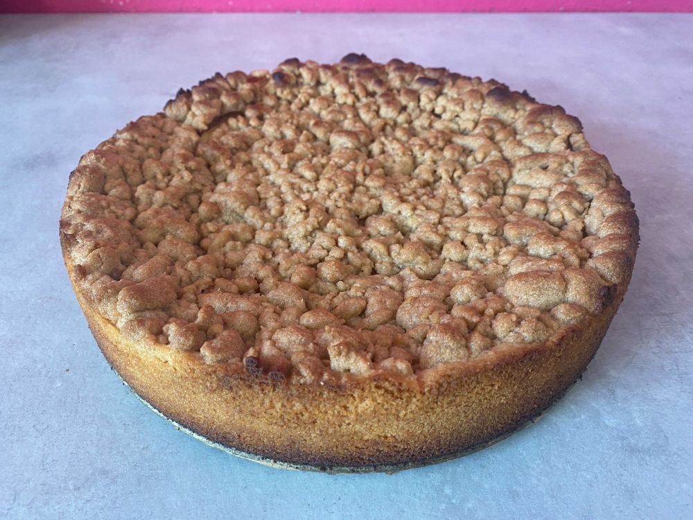 Ein frisch gebackener runder Apfelkuchen steht auf einer grauen Arbeitsplatte. Der Kuchen ist noch nicht angeschnitten, man sieht daher nur den Mürbeteigrand und die Streuseldecke.