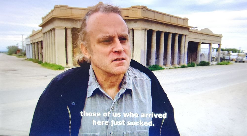 Brad Dourif standing in front of abandoned stores wearing a light blue shirt and dark blue hoodie from Warner Herzig's "The Wild Blue Yonder".