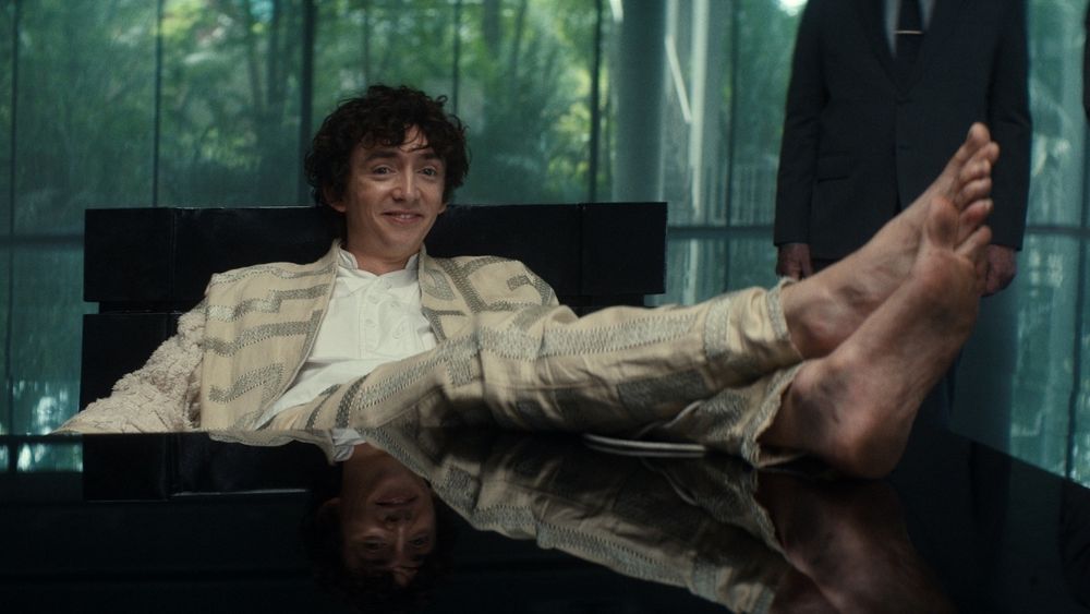 Samuel Blenkin as Kid Kavalier in Alien: Earth. Uncombed messy hair, smirking and wearing pajama-like pants, barefeet set on a conference table during legal proceedings