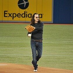 Geddy Lee in blue jeans and long sleeve black shirt throwing out the first pitch of a Toronto game