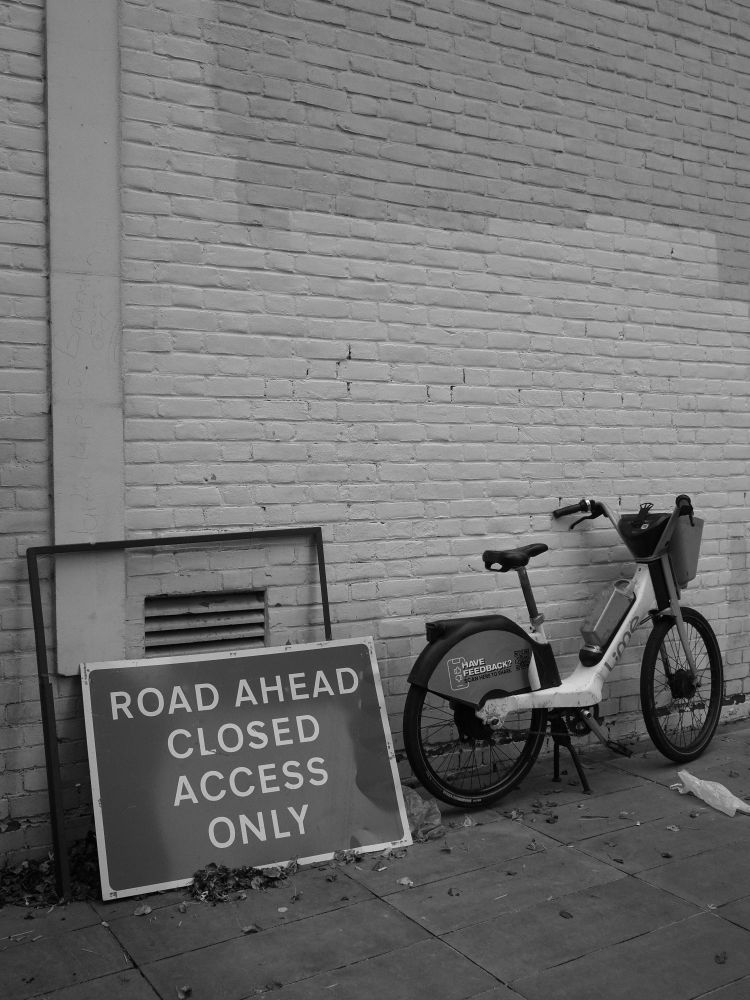 Monochrome photograph. A sign leaning against a whitewashed wall reads, 'Road ahead closed access only'. A Lime bike leans against the wall next to it.