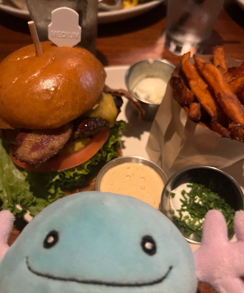 A delicious plate of a bacon burger with a side of sweet potato fries, as well as various condiments on the side. My beloved Wolpy the Wooper plush is photobombing.