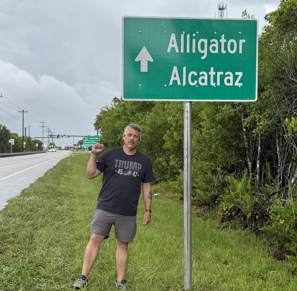 A man in a Trump shirt standing next to an Alligator Alcatraz sign