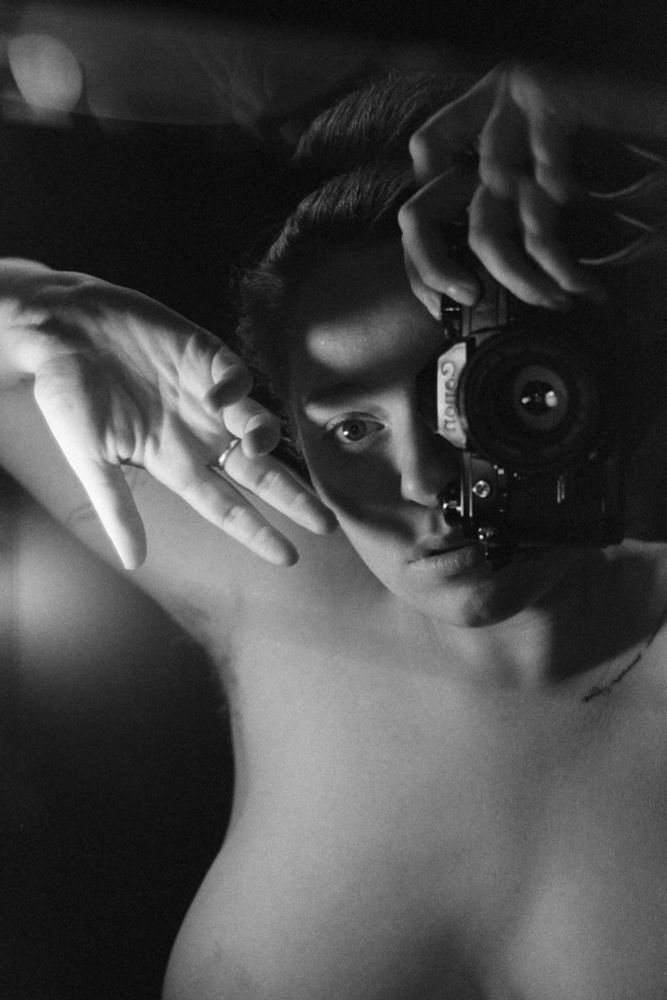 a 35mm film black and white self portrait image of a woman. there is a soft white light coming from the side of the frame, which she holds her hand up to, casting an intense shadow across her face. She is using a Canon ae1 to take the photo, staring into the mirror. Photo by Charlotte J Mack 
