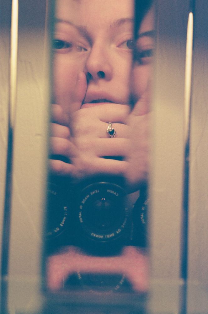 a 35mm color photograph taken in a narrow mirror. the photographer holds the camera below their face, with their hand across the top of the camera resting on their chin. the edges of the mirror reflect the image thrice, so the photographers face is mimicked upon itself. taken by Charlotte j mack 