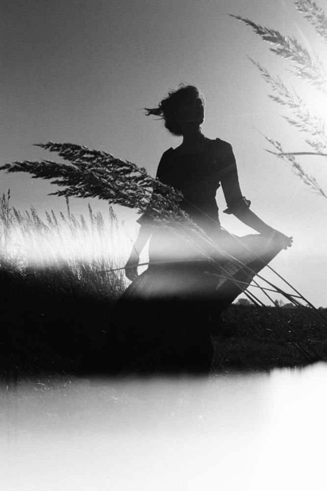 a 35mm black and white image. The bottom quarter of the image is pure white, burned from being the first of the roll. In the foreground, stocks of wheat blow diagonal from the right corner. A woman stands behind the wheat, holding her dress, hair up and head turned in profile. She is in silhouette. photo by Charlotte j mack 