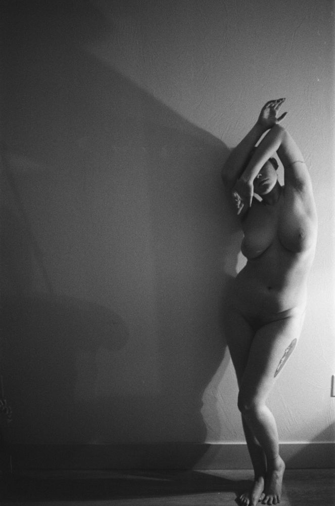 a 35mm black and white film photo. a woman stands nude on the right side is the frame against a blank wall. there is storm light from the side illuminating her body. she leans against the wall with arms across her face. self portrait by charlotte j mack 