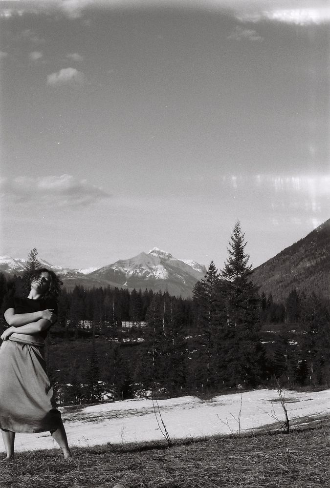 a black and white film image of a woman on a hillside, with pine trees behind her and a bold mountain in the far background. She is wearing a long skirt and a black shirt. She is standing at the left edge of the image, with her arms wrapped around herself, leaning back with eyes closed and face angled toward the light. 35mm self portrait taken by Charlotte j mack. 
