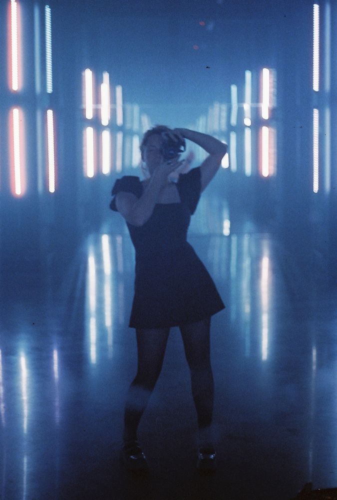 A 35mm color film photograph. Taken in a mirror, a woman in a short black dress with black nylons holds a canon ae1 in front of her face. behind her, a wide space with blue and orange lights stretches far behind her. taken by Charlotte j mack