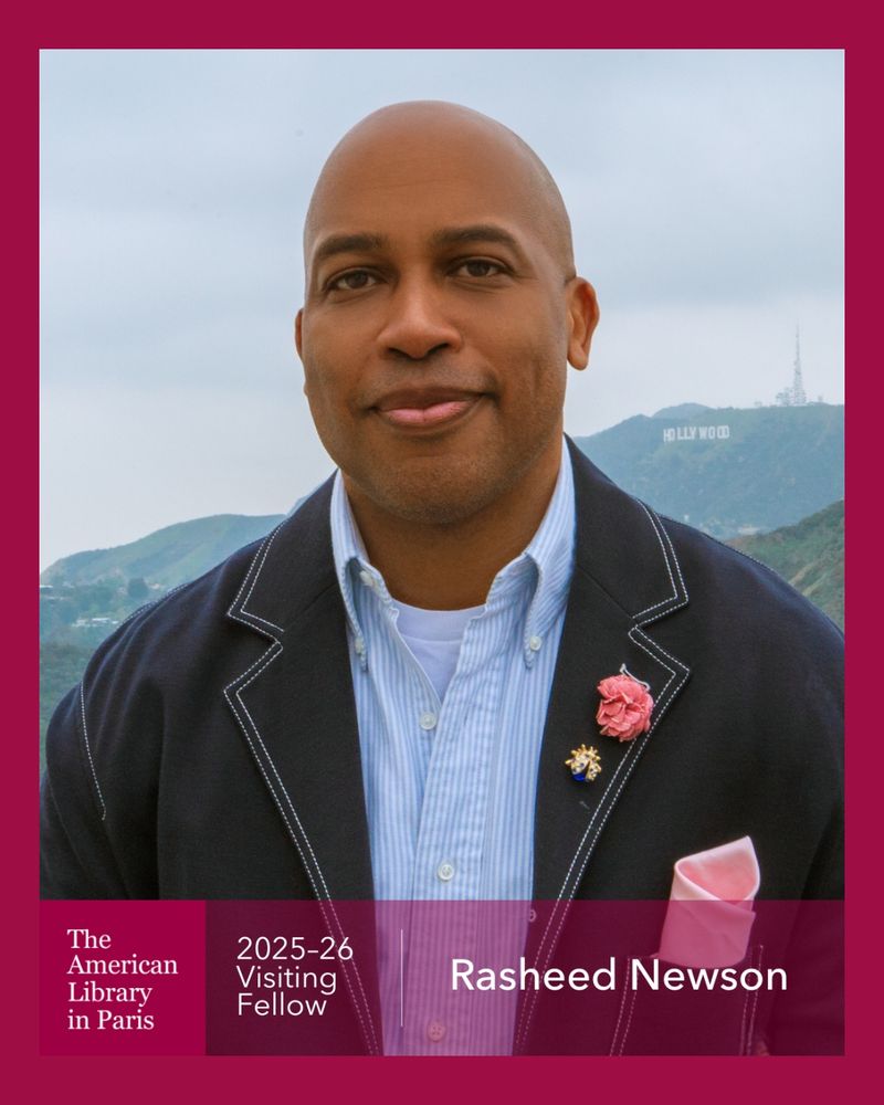 Headshot of Visiting Fellow Rasheed Newson. He is looking at the camera in a blue button down and navy blue jacket with a pink flower pin and gold bug pin on it. You can see the "Hollywood" sign in Los Angeles in the background. The sky is gray. The bottom of the image has The American Library in Paris logo with the text "2025-26 Visiting Fellow Rasheed Newson"