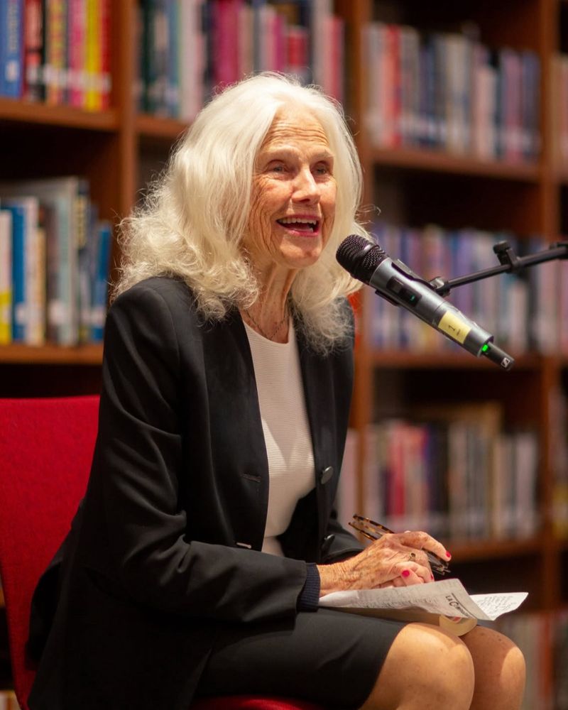 Image of author Sheila Kohler sitting in a red chair, speaking into a microphone with a line of bookshelves behind her.