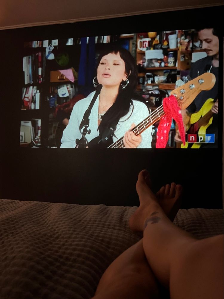 Saya Gray’s Tiny Desk concert projected on a wall, Blake’s legs on a bed in the foreground 