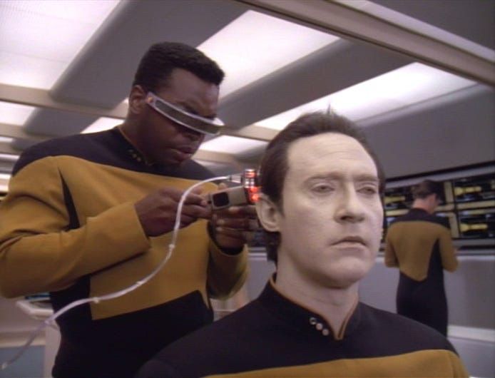 Geordi is examining data holding a diagnostic tool 