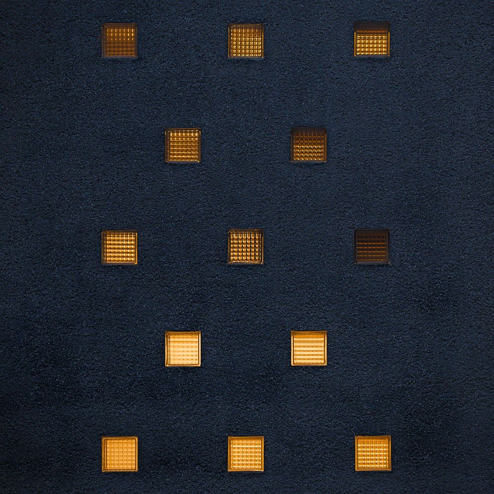 Ein quadratisches Foto. Zu sehen ist eine dunkelblaue raue Hausfassade, darin orange leuchtende Glassteine.