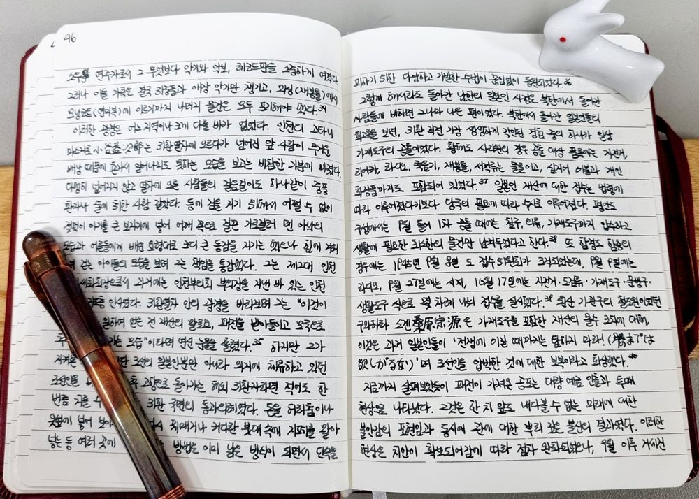 필사 중인 공책. 양옆으로 펼쳐진 종이 양면에 검은 잉크로 적은 글씨가 빼곡하다. 좌측 하단에는 검게 녹이 올라온 구리 만년필이, 우측 상단에는 하얀 토끼 장식품이 공책을 누르고 있다.