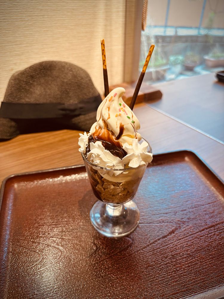 お盆の上に、チョコサンデーが乗った小さい足つきグラス。グラスの中には下からコーンフレーク、バニラソフトクリーム、生クリーム、チョコシロップ、チョコスプレー、ポッキーが2本刺さっている。