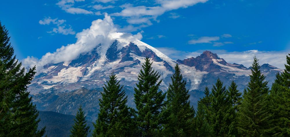 Photo of Mt Rainier on a mostly clear day. Clouds are blowing over the summit and trees fill the foreground. 