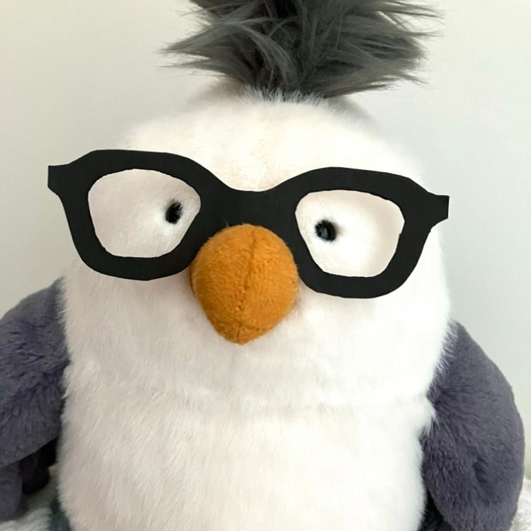A photo of a seagull plushie named John Gulliver with thick paper glasses on orange beak and a tuff of grey fur on the head. The soft fur toy is starting directly at the viewer, smiling.