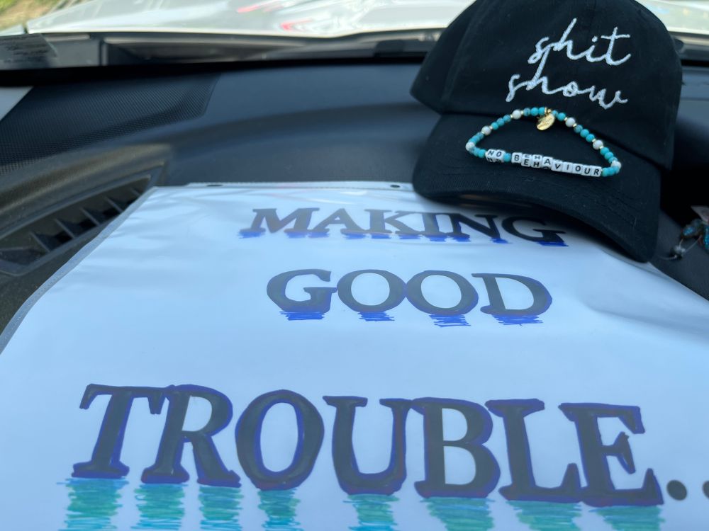 Sign reading Making Good Trouble, a hat reading shit show, and a bracelet reading no behaviour. 