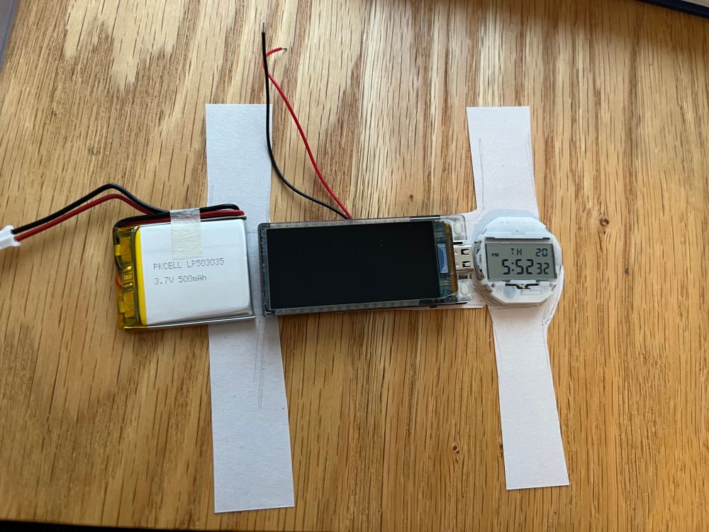 An image of a watch with two paper straps, made up of the body of a casio digital watch, a microcontroller, and a lipo battery
