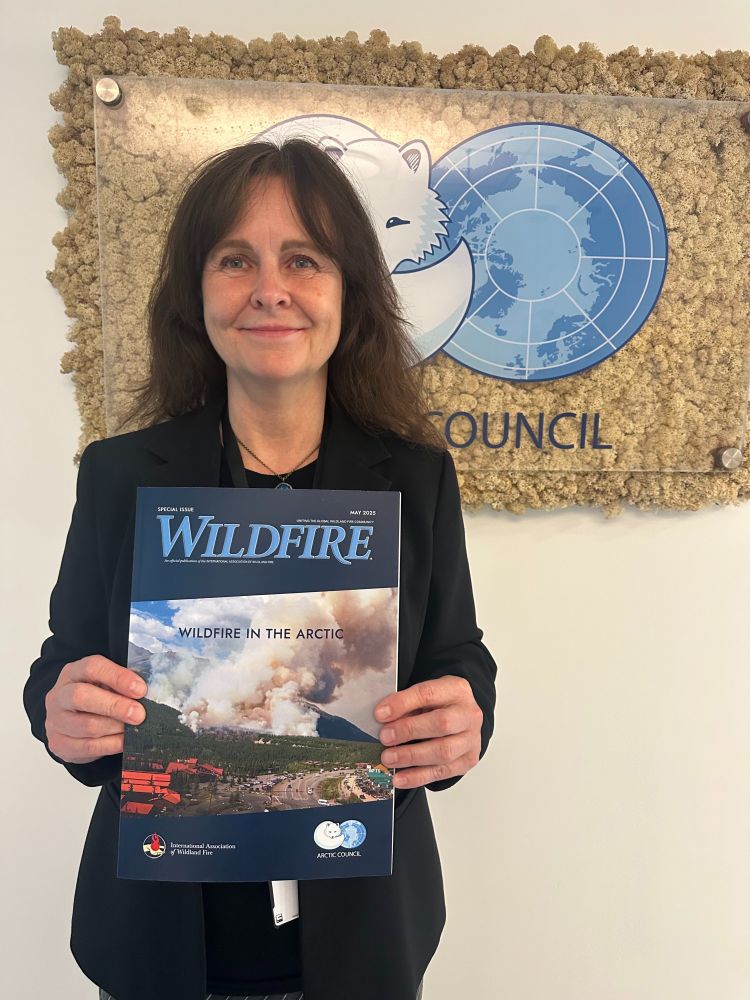 person wearing black blazer holds a copy of the Arctic issue of Wildfire magazine and smiles. Behind her is a plaque displaying the Arctic Council’s logo, an Arctic fox next to a circumpolar map.