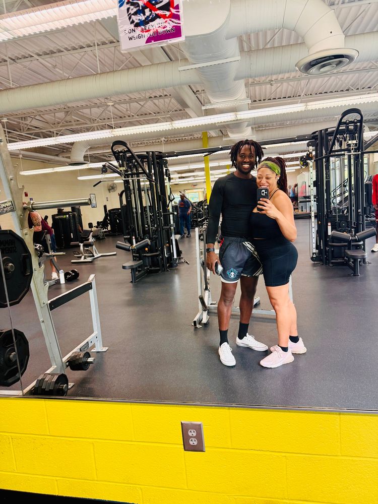 A Black woman and man with locs taking gym selfie in a gym mirror after their individual workouts