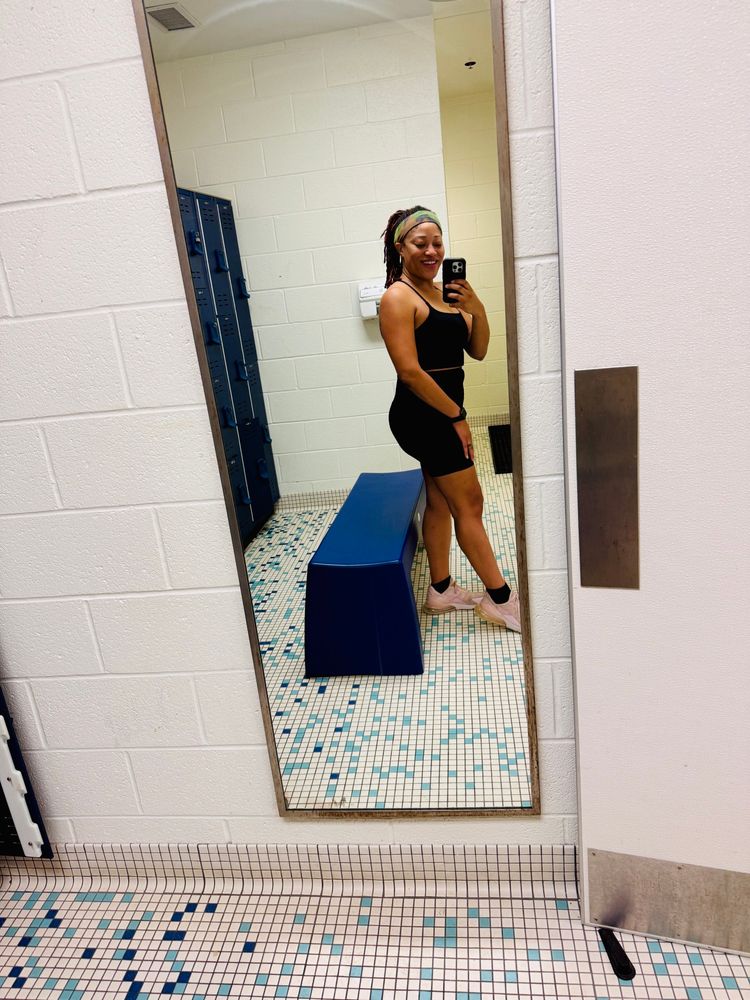 A Black woman with locs taking gym selfie in a locker room mirror