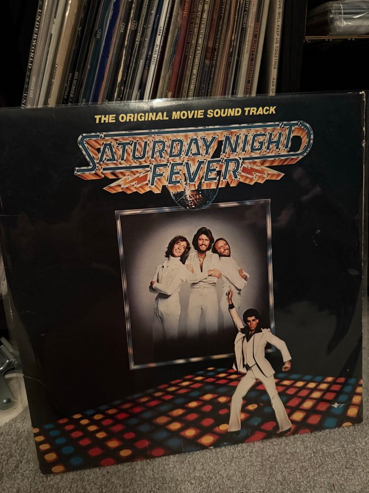 The jacket of the Saturday Night Fever soundtrack LP, leaning against my shelf which has a number of other LPs. The front of the jacket has a photo of John Travolta in his iconic pose on the dance floor, one foot in front of the other in a wide stance while his hip is thrust back. His left arm is down and behind him while his right arm is up in the air, pointing. He is wearing a whole suit and vest with a dark shirt. Wide lapels and collars. Totally stylin. The dance floor is square with different coloured lights underneath. The background in the centre is a photo of the Gibbs brothers (aka the Bee Gees), Robin on the left, Maurice on the right, and Barry in the center. All smiling, all wearing white. 