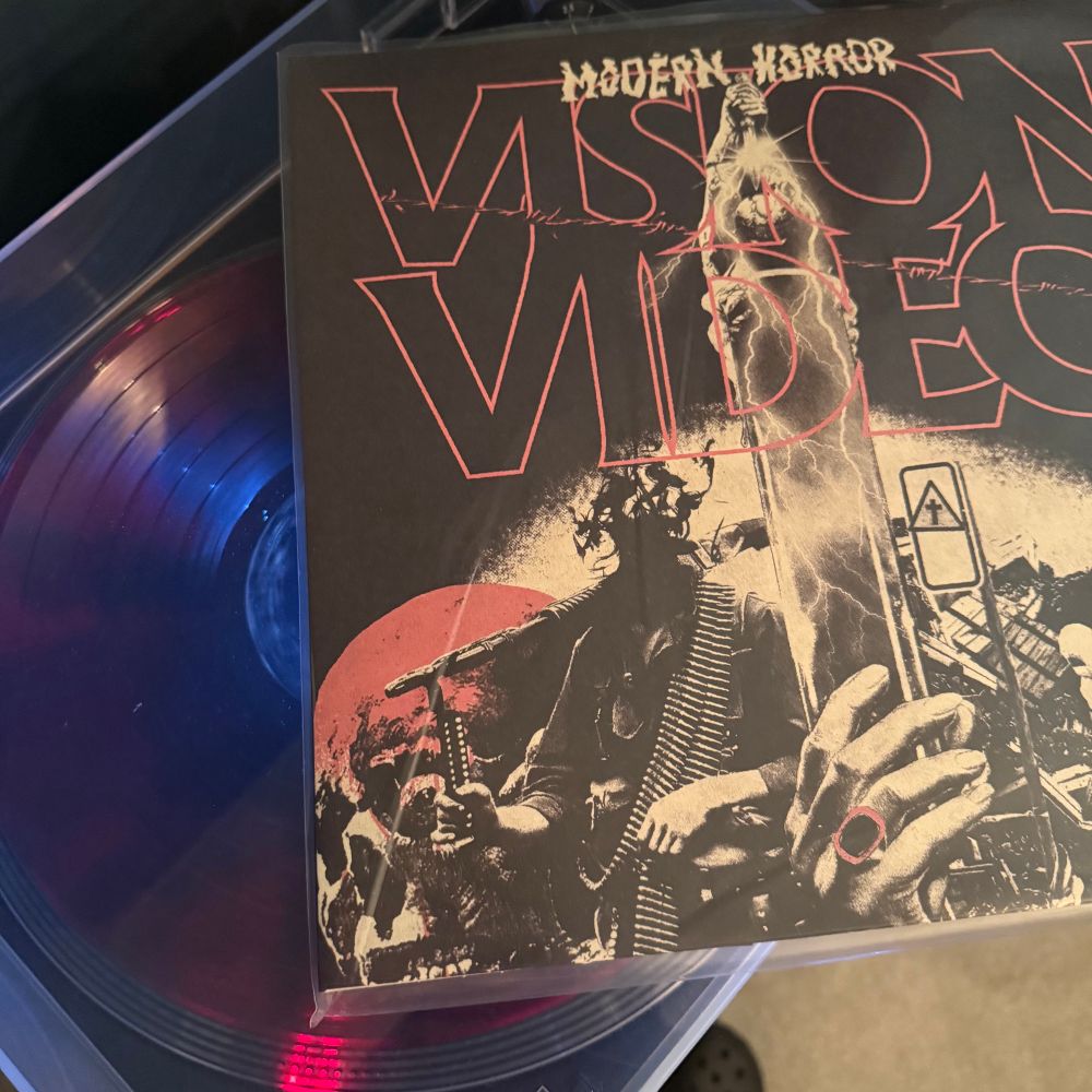 The jacket of Vision Video's Modern Horror LP lies on the lid of my turntable with the LP spinning underneath. 

The art is a collage of a couple of skulls on the right and left-hand side (each with one eyeball in a socket), a soldier with a machine gun across their shoulder along with at least one bandolier of ammunition. Their head is obscured by smoke to the point it seems they don't have a head. Someone in a hazmat suit with what looks to be rubble and a sign that has a triangle and in it is a "Christian" cross. In the centre, a hand holding something that seems metallic with lightning bolts reflected on its surface. This leads up to a person holding a dagger above their head with both hands and the blade pointing down. The blade gleams and makes it difficult to discern. 

Across the top half of the jacket is the "vision video" text logo with a strand of barbed wire across from the top left to the bottom right. Just above that is the title of the record "Modern Horror" in a ragged typeface.