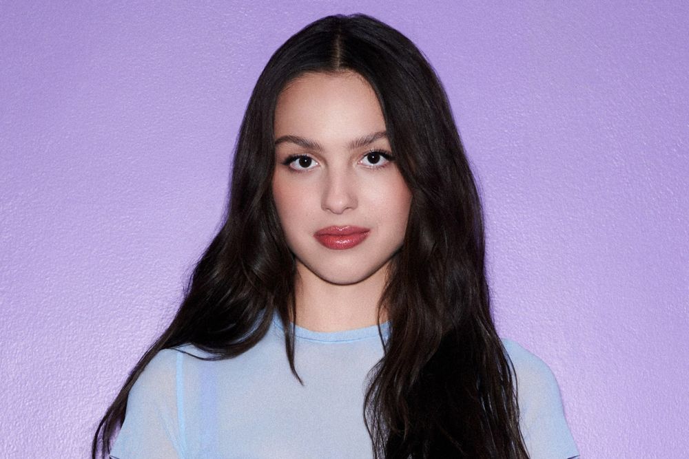 A headshot photo of Olivia Rodrigo. She is a young woman with light skin (she is "half-Filipina" according to Wikipedia), long brown hair, and dark eyes, wearing a blue blouse.