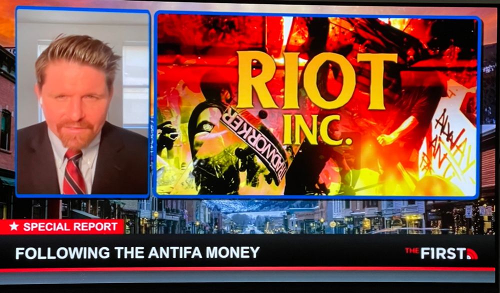 White douchebag from last photo next a large graphic that is an abstract collage of what’s supposed to be riots, with protestors, signs, fire, it’s mostly red and black. Text in yellow reads “riot inc”