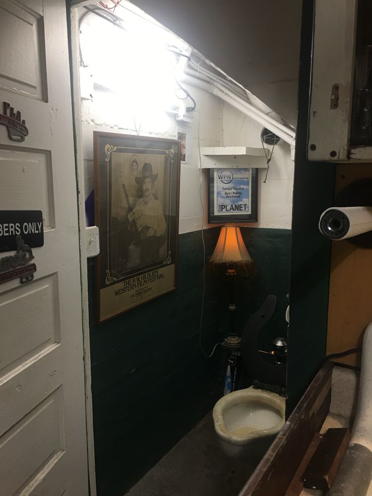 Looking into a bathroom with the door open, there’s an antique lamp, half green walls, some wires and other items that would make it seem less than safe. The toilet itself has a nasty grime of filth on it. On the wall, a poster for a western film festival and in a corner, a sign from a Weekly Planet award naming it the best public restroom in Tampa in 2001. 